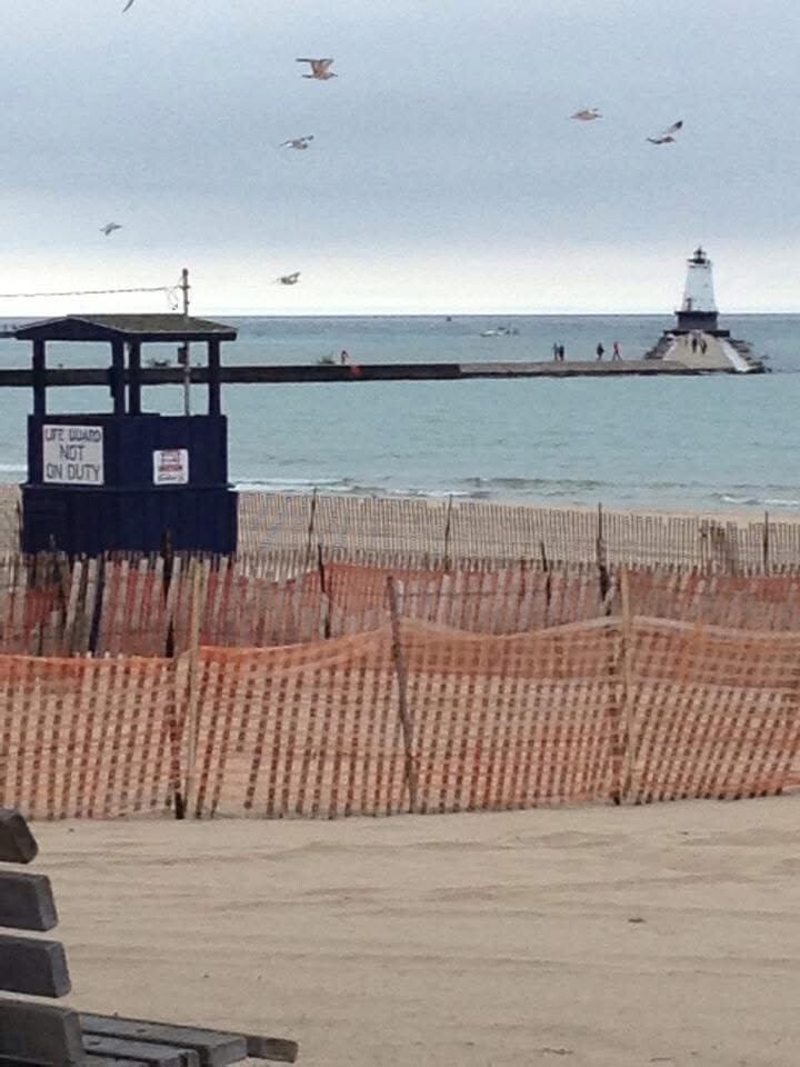 Lifeguard stand marked "NOT ON DUTY" on a beach with sand fencing, overlooking a breakwater and lighthouse across the water.