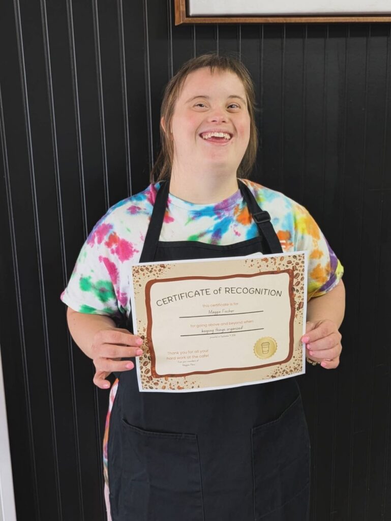 A person wearing a colorful tie-dye shirt and black apron holds a Certificate of Recognition in front of a dark striped wall.