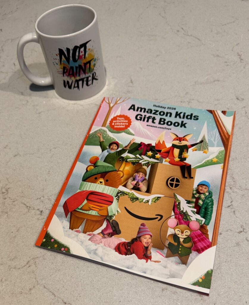 A white mug with "Not Paint Water" text sits next to the Holiday 2026 Amazon Kids Gift Book on a concrete surface.
