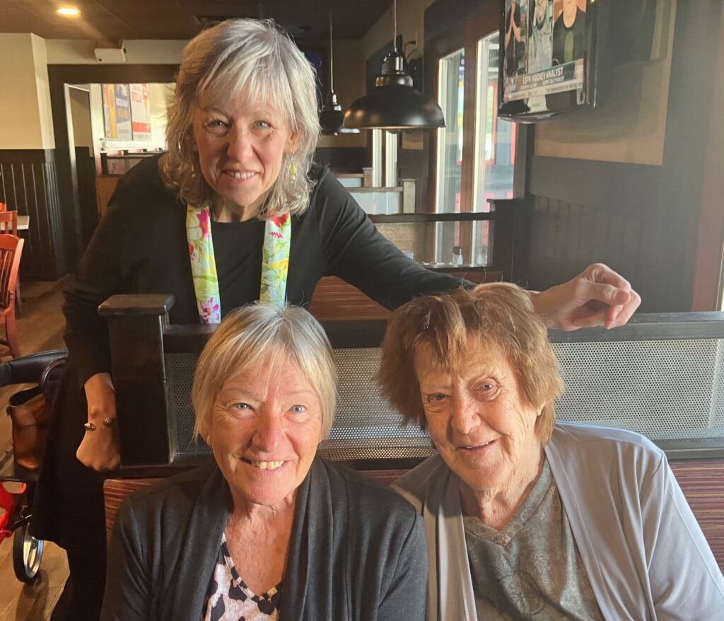 Three smiling women pose together at a counter in a modern café with pendant lighting and large windows in the background.